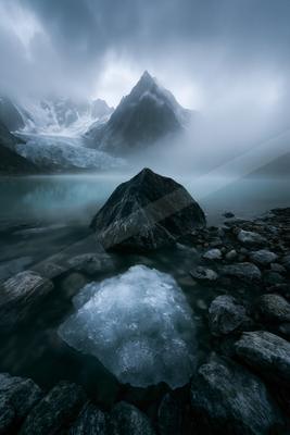 A serene glacier lake surrounded by rocky shores, featuring a floating iceberg and a majestic snow-capped mountain peak in the background.