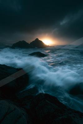 Powerful ocean waves with white foam crash against dark rocks in the foreground while the sun sets behind a silhouetted rocky island under a moody, cloudy sky.