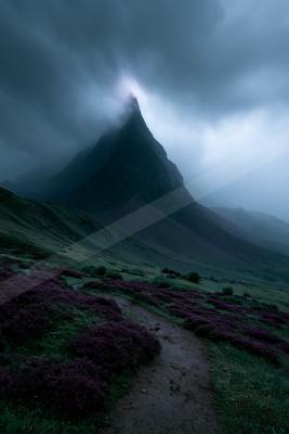 A steep rocky mountain peak shrouded in thick fog and low clouds, with a foreground of vibrant purple heather and a winding dirt path through green hills under 