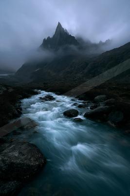 A fast-flowing river with white water rushes over rocks through a rugged mountain valley shrouded in thick fog, with a sharp rocky peak rising in the background
