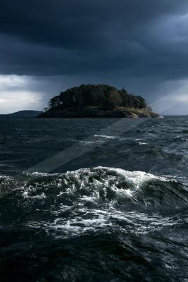 A small forested island rises from turbulent ocean waters under a moody sky filled with dark storm clouds.