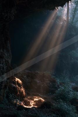 Dramatic sunbeams streaming into a dark rocky cave entrance surrounded by lush green forest foliage, illuminating the cave floor with golden light.