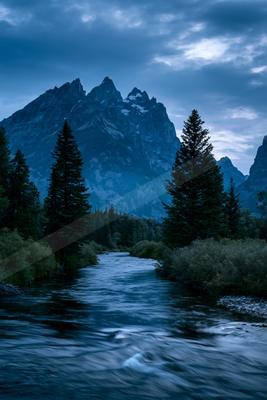 A fast-flowing river winds through a dense evergreen forest toward jagged, snow-dusted mountain peaks under a dramatic cloudy sky.