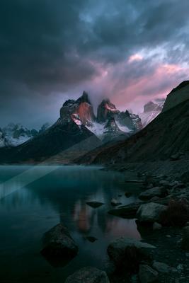 A majestic snow-capped mountain peak is illuminated by the soft glow of twilight, casting a serene reflection on a calm lake surrounded by rugged terrain.