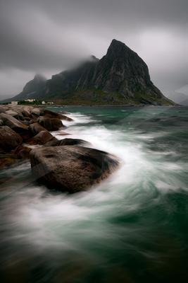 Turquoise ocean waves crash against large boulders on a rocky shore, with a steep, mist-covered mountain rising in the background under an overcast sky.