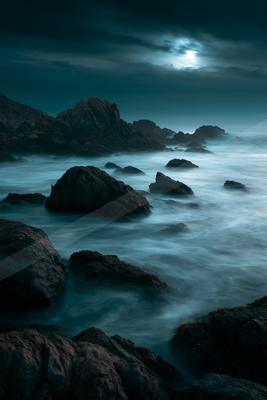 Dark rocky shoreline with smooth misty water flowing around boulders under a cloudy night sky with moonlight breaking through