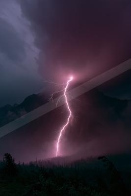 A bright pink lightning bolt strikes down from dark storm clouds onto a forested mountain peak, illuminating the dramatic sky in shades of purple and magenta.