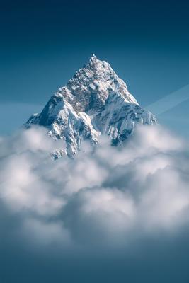 A sharp, snow-covered mountain peak emerges dramatically from a dense layer of white clouds under a clear deep blue sky.