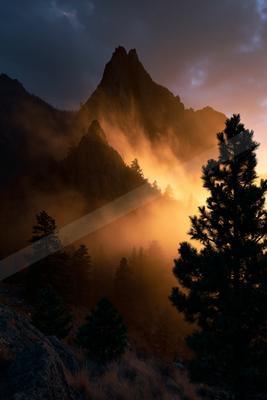 A rugged mountain peak is silhouetted against a vibrant orange sunset, with mist swirling through the forest below.