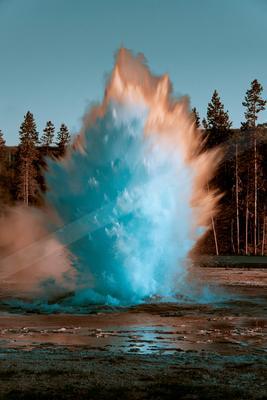 A powerful geyser erupts with a massive plume of bright blue and orange water and steam, set against a backdrop of tall pine trees under a clear blue sky.