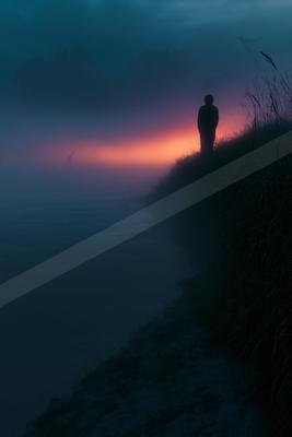 A silhouetted person stands on a grassy bank overlooking a misty lake at dusk, with a vibrant orange and purple glow on the horizon and birds flying in the fogg